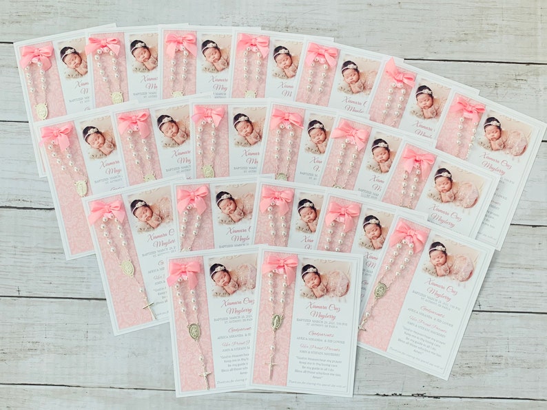 May include: Pink and white cards with a photo of a baby and a small rosary with a silver charm. The cards have the text "Anamara Grace" and "Baptized into the Love of God" on them.