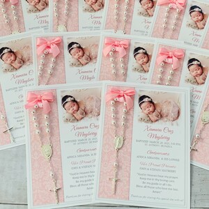 May include: Pink and white baptism favor cards with a photo of a baby girl, a silver rosary, and the text "Xiomara Ouz Mayberry Baptized March 24, 2001, 3:00 PM St. Anthony de Padua Godparents Africa Miranda & Ed Lowery Her Proud Parents Jorn & Stefano Mayberry "God in Heaven hear my prayer Keep me in thy loving care Be my guide in all I do. Bless all those who love me too. Amen" Thank you for sharing this special day with us!"