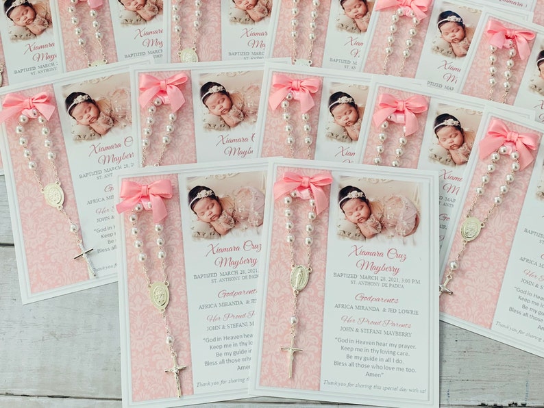 May include: Pink and white baptism favor cards with a photo of a baby girl, a silver rosary, and the text "Xiomara Ouz Mayberry Baptized March 24, 2001, 3:00 PM St. Anthony de Padua Godparents Africa Miranda & Ed Lowery Her Proud Parents Jorn & Stefano Mayberry "God in Heaven hear my prayer Keep me in thy loving care Be my guide in all I do. Bless all those who love me too. Amen" Thank you for sharing this special day with us!"