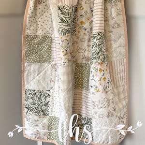 Puede incluir: Un edredón patchwork con una variedad de patrones, incluyendo lunares, rayas y diseños florales. El edredón presenta una paleta de colores suaves de blanco, verde y beige, con un borde rosa. El texto "Little Homemade Shop" es visible.