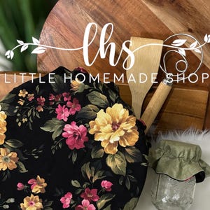 Può includere: Un copripentola rotondo in tessuto nero con un motivo floreale in tonalità di rosa, giallo e verde. Il copripentola è su un tagliere di legno con un cucchiaio di legno e una frusta. Il testo "lhs LITTLE HOMEMADE SHOP" è visibile sul tagliere.