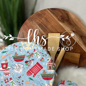 May include: A set of two blue and white fabric bowl covers with a gingerbread cookie and Christmas baking theme. The covers feature a variety of festive designs, including gingerbread houses, candy canes, and snowflakes. The text "Oh Christmas Cookies" is printed on the covers.