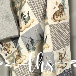 Könnte beinhalten: Eine Babydecke mit Patchwork-Muster, das verschiedene Drachenmotive zeigt. Die Decke ist aus weichem, plüschem Stoff gefertigt und perfekt zum Kuscheln. Die Decke ist grau, blau und weiß mit einem braunen Drachenmuster. Die Decke trägt den Text "Little Homemade Shop" unten.