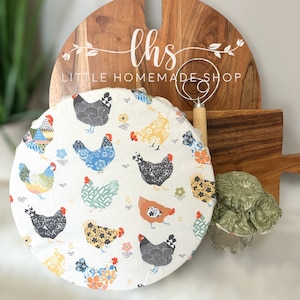 Puede incluir: Una cubierta de cocina redonda de tela blanca con estampado de pollo. La cubierta está colgada de una tabla de cortar de madera con el texto "lhs LITTLE HOMEMADE SHOP" en ella.