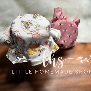 May include: Two fabric bee-themed jar covers. One is white with gold accents and a honeycomb pattern. The other is pink with a smaller bee pattern. The covers are on a wooden surface. The text "Little Homemade Shop" is visible below the covers.