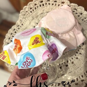 May include: A white fabric jar lid with a pink ruffle and a print of colourful candy hearts with messages like "Kiss Me", "Love You", and "Hugs".