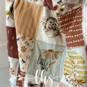 Puede incluir: Un edredón de retazos con cuadrados que presentan diseños de animales del bosque y florales. El edredón incluye imágenes de un erizo, un oso y margaritas, con el texto "little one" y "lhs homemade shop". La paleta de colores incluye tonos de marrón, crema y verde.