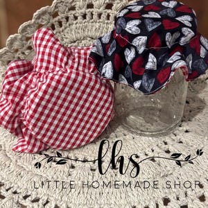 May include: Two fabric jar covers, one with a red and white gingham pattern and the other with a black background and white and red hearts. The covers are sitting on a crocheted doily with the text "lhs LITTLE HOMEMADE SHOP".