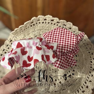 May include: A glass jar with a red and white gingham fabric lid and a white fabric lid with pink and red hearts. The jar is sitting on a crocheted doily. The text "lhs LITTLE HOMEMADE SHOP" is printed on the jar.