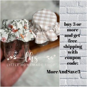 May include: Two glass jars with fabric lids. One lid is a white and brown gingham pattern and the other is a floral pattern with pink, orange, and green flowers. The jars are on a wooden surface. The text "ths LITTLE HOMEMADE SHOP" is below the jars. The text "buy 3 or more and get free shipping with coupon code: MoreAndSave3" is on a white brick background.
