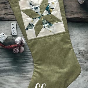 May include: A green fabric Christmas stocking with a white star-shaped patchwork design. The stocking is decorated with a white floral pattern and has the text "lhs LITTLE HOMEMADE SHOP" at the bottom.