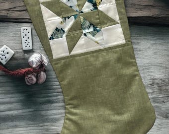 Op de afbeelding: Een groene stoffen kerstsok met een wit, stervormig patchwork-ontwerp. De sok is versierd met een wit bloemenpatroon en heeft de tekst "lhs LITTLE HOMEMADE SHOP" aan de onderkant.