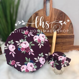 May include: Two burgundy fabric covers with a floral print of pink and white roses and green leaves. The covers are for jars and bowls. The larger cover is round and the smaller cover has a ruffled edge. The covers are on a white surface with a wooden chopping board in the background. The chopping board has the text "lhs LITTLE HOMEMADE SHOP" on it.