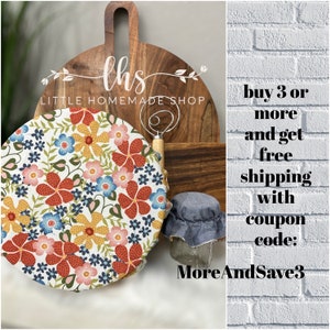 May include: A round, white fabric cover with a floral pattern in shades of red, yellow, blue, and pink. The fabric cover is on a wooden chopping board with a sign that reads "ths LITTLE HOMEMADE SHOP". The image also includes a white brick wall with text that reads "buy 3 or more and get free shipping with coupon code: MoreAndSave3".