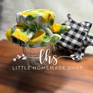 May include: A glass jar with a lemon-print fabric lid and a black and white gingham lid. The jar has the text "lhs LITTLE HOMEMADE SHOP". The lids are on a wooden surface.
