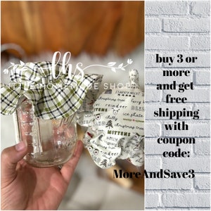 May include: A glass jar with a green and white plaid fabric lid. The fabric has a Christmas theme with words like "Christmas tree", "reindeer", "mittens", and "holly". The text "buy 3 or more and get free shipping with coupon code: MoreAndSave3" is visible on a white brick background.