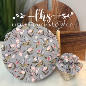 May include: Two grey fabric covers with a floral pattern in shades of pink and brown. The covers are for jars or bowls. The text "lhs LITTLE HOMEMADE SHOP" is on a wooden chopping board in the background.