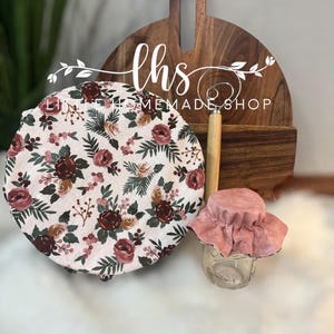 May include: A set of two floral fabric food covers. The larger cover features a pink and green floral pattern with the text "Little Home Made Shop" and the smaller cover is a solid pink colour.