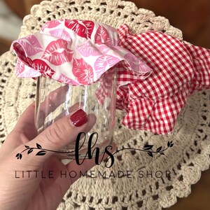 May include: Two fabric jar covers. One is white with pink and red lips printed on it. The other is red and white gingham. The covers are on a glass jar. The jar is sitting on a crocheted doily. The text "Little Homemade Shop" is visible in the image.