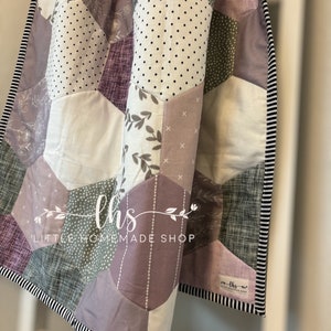 May include: A purple, green, and white patchwork quilt with a hexagonal pattern. The quilt has a black and white striped border and a label that reads "lhs little homemade shop".