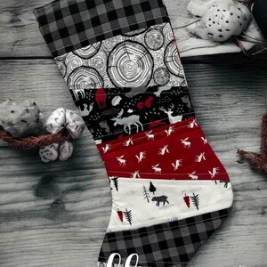 Op de afbeelding: Een rode, witte, zwarte en grijze kerstsok met een patchwork-ontwerp met afbeeldingen van bosdieren, bomen en boomringen. De sok heeft een lus aan de bovenkant om hem op te hangen. De sok is gelabeld met "Little Homemade Shop".