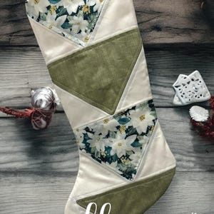 May include: A white Christmas stocking with a green, white, and floral pattern. The stocking is made of fabric and has a loop at the top for hanging. The stocking is on a wooden background. The text "lhs LITTLE HOMEMADE SHOP" is at the bottom of the image.