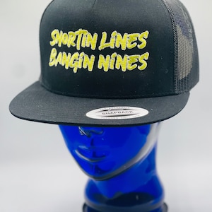 May include: Black and yellow embroidered trucker hat with the text "Shortin Lines Bangin Nines" on the front. The hat has a black mesh back and a black snap closure.