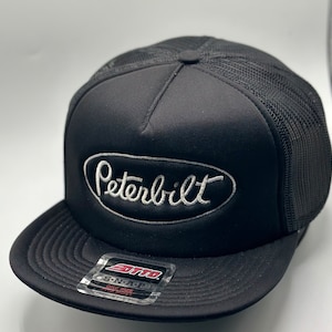 Op de afbeelding: Zwarte truckercap met een wit geborduurd Peterbilt-logo. De pet heeft een mesh achterkant en een zwarte drukknopsluiting.