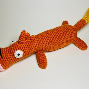 Puede incluir: Un animal de peluche tejido a crochet con forma de perro de cuerpo largo con nariz rosa, ojos blancos y cola amarilla.