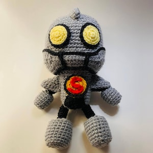 May include: A gray crocheted robot toy with yellow eyes, a red 'S' on its chest, and black legs.