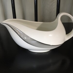 May include: White porcelain gravy boat with a black and silver design. The boat has a curved handle.