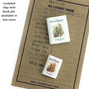 Może przedstawiać: Miniaturowa szpilka do książek z gliny przedstawiająca "Opowieść o Króliku Peterze" Beatrix Potter. Szpilka jest repliką okładki książki i zawiera tytuł, autora i ilustracje. Szpilka jest dostępna w dwóch rozmiarach. Szpilka jest prezentowana na brązowej kartce papieru z listą dat i tekstem "The Literary Charm".