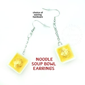 May include: A pair of noodle soup bowl earrings. The earrings are made of white ceramic with a yellow glaze and feature a realistic depiction of noodles in a bowl. The earrings are attached to silver chains and hooks.