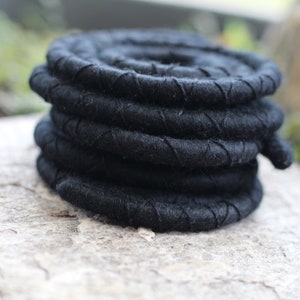 May include: A coiled black rope made from a thick, textured material. The rope is tightly wound into a circular shape, with visible stitching along the edges. The rope is resting on a light-coloured, textured surface.