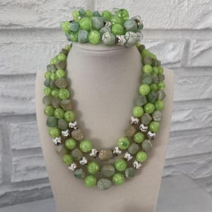May include: A three-strand necklace, bracelet, and matching earrings set. The jewelry features green and gray beads with white accents. The necklace and bracelet are displayed on a mannequin. The earrings are on a clear stand.
