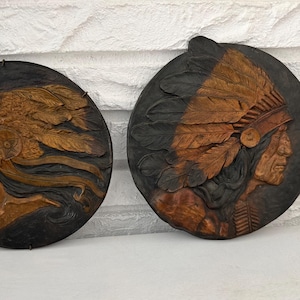 May include: Two circular wall plaques featuring detailed relief carvings of a Native American chief in profile. The carvings are in shades of brown and tan against a dark background, showcasing the chief's headdress and facial features.