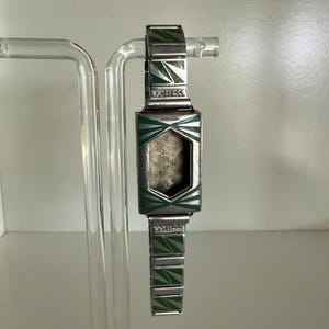 Pode incluir: Uma caixa de relógio prateada com pulseira de elos apresentando detalhes geométricos em esmalte verde. A cabeça retangular do relógio tem um recorte central e a pulseira de metal exibe um padrão triangular repetitivo. A palavra Diehl é visível.