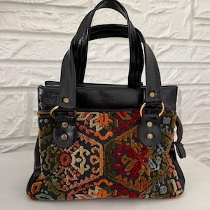 May include: A handbag featuring a black patent leather top section and handles with gold-tone metal rivets. The main body displays a multi-colored woven tapestry pattern with geometric shapes in shades of red, orange, green, and blue on a black base.