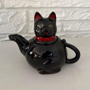 May include: Black ceramic cat teapot with a red bow tie. The teapot has green eyes and red inner ears. The spout and handle are curved. The teapot is sitting on a white surface.