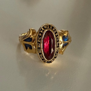 May include: A gold-colored ring with a large, oval, red gemstone at the center. The ring has a black inscription around the gemstone and blue accents on the sides. The ring is likely a class ring or a similar commemorative piece.
