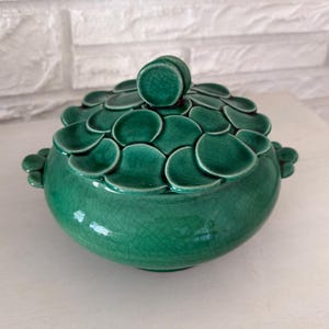 May include: A round, emerald green ceramic container with a lid. The lid is adorned with overlapping, petal-shaped elements. The container has two small handles on either side and a textured, crackled surface.