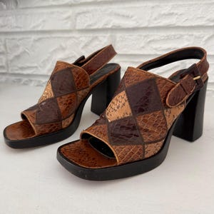 May include: A pair of open-toe sandals featuring a brown and tan snakeskin-patterned patchwork design in a diamond motif. The sandals have a thick black platform sole, a chunky black block heel, and an adjustable slingback strap with a buckle.