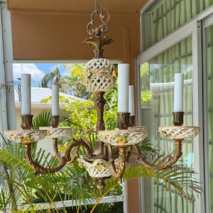 May include: An ornate, vintage chandelier with a bronze finish. It features six candle holders with white candles, and decorative ceramic accents with floral patterns. The chandelier is suspended from a decorative ceiling mount.