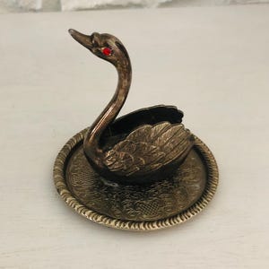 Puede incluir: Figura de cisne plateado con ojos de gema roja, posada sobre una base redonda decorativa. La base tiene un patrón en relieve y un borde con cuentas. Las alas del cisne son detalladas, y el diseño es vintage.