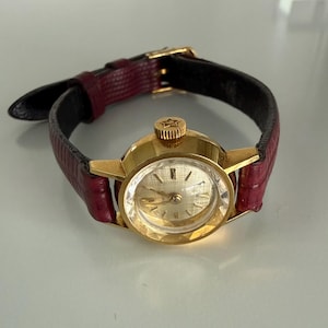 May include: A gold-tone wristwatch featuring a round, faceted crystal face with a textured silver dial and gold-tone markers. The watch has a dark red, textured leather strap with a gold-tone buckle and a crown embossed with a star symbol.