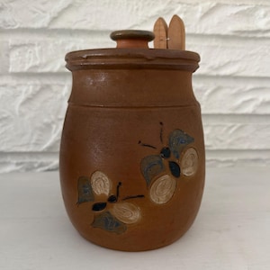 May include: A brown ceramic jar with a lid and two wooden spoons. The jar is decorated with two butterfly designs in blue, tan, and white. The jar is sitting on a white surface.