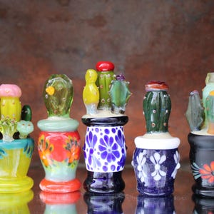 May include: Five colorful glass cactus bottle stoppers with different designs. The stoppers are in various shades of green, yellow, blue, red, and black. Each stopper features a unique cactus design with intricate details.