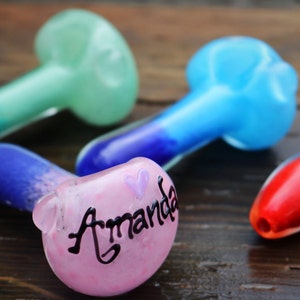 May include: Four glass pipes in different colors, including pink, blue, green, and red. The pink pipe has the name "Amanda" written on it with a small purple heart.