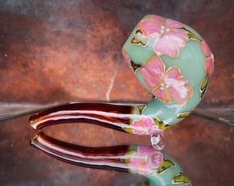 May include: A glass pipe with a light green bowl and a brown stem. The bowl is decorated with pink flowers.