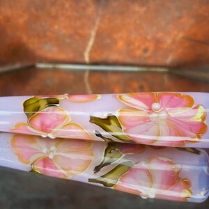 May include: A light purple glass bead with a floral design. The bead features pink flowers with white centers and green leaves.